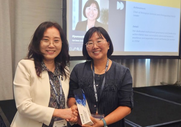Cal State LA Biological Sciences chair Hyunsook Park smiles while holding her 2026 Outstanding Woman Leadership Award and shaking hands with another attendee at the UKC 2026 Women's Forum. A screen behind them displays her photo and award recognition.