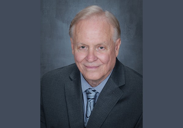 Portrait of Cal State LA Professor of Hydrogeology Barry Hibbs wearing a dark suit and patterned tie against a gray-blue background.