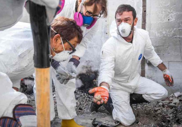 A masked archaeologist kneels in the rubble of a burned home, carefully holding a small, recovered object while colleagues look on.