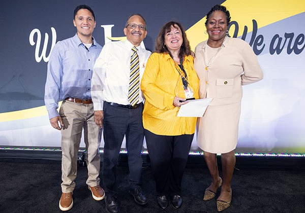 Four individuals stand together on a stage during an employee recognition event. The award recipient, Karin Elliott Brown, wearing a bright yellow jacket, holds a recognition award and certificate while posing with President Eanes and her family. A black and gold backdrop behind them features the phrase "We Are" as part of the event branding.