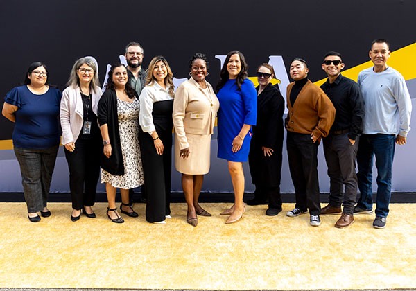 A group of ten employees stands together at the employee recognition event, smiling toward the camera. The group poses in front of a black and gold event backdrop, with two individuals standing at the center and colleagues gathered on either side.