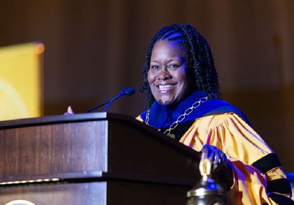 Cal State LA President stands at a podium in academic regalia, smiling while speaking during a ceremony.