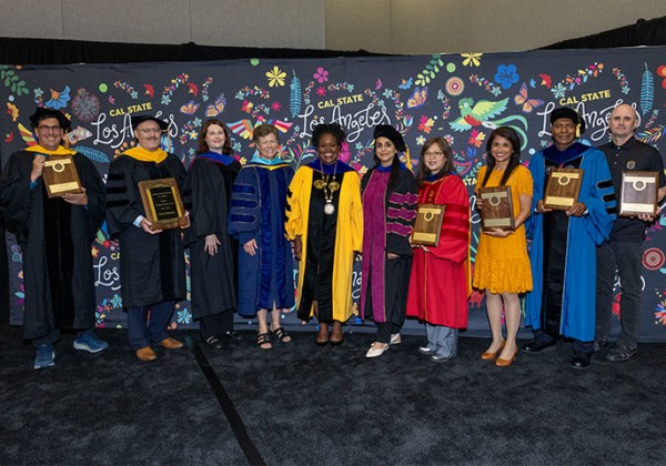 A group of ten Cal State LA faculty and campus leaders stand together on a stage in academic regalia, with several individuals holding award plaques. They pose in front of a black backdrop featuring colorful floral artwork and repeated Cal State Los Angeles logos during a faculty recognition ceremony.