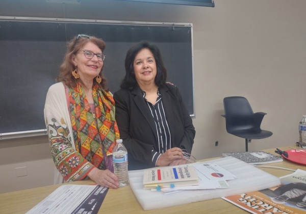 Two speakers stand behind a table in a classroom, with books and papers laid out in front of them and a chalkboard behind them at Cal State LA.