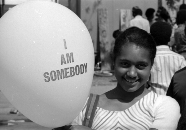 A young girl smiles while holding a balloon that reads “I AM SOMEBODY.” She stands outdoors among a crowd, wearing a striped top, with trees and people softly blurred in the background of this black-and-white photograph.
