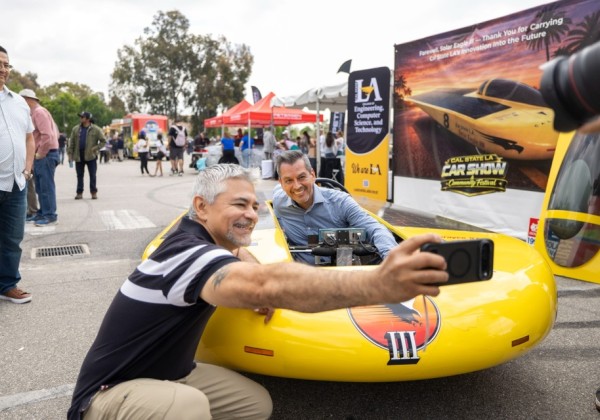 Two attendees take a selfie while seated in a yellow solar-powered vehicle at a Cal State LA car show event.