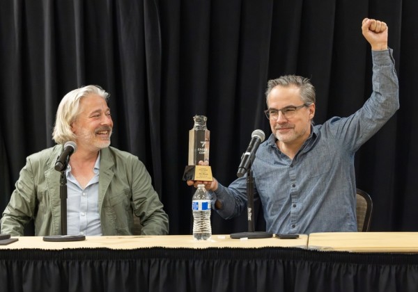 Two panelists at Cal State LA’s Eagle-Con sit at a table with microphones. One smiles while the other holds up an award trophy with a raised fist.