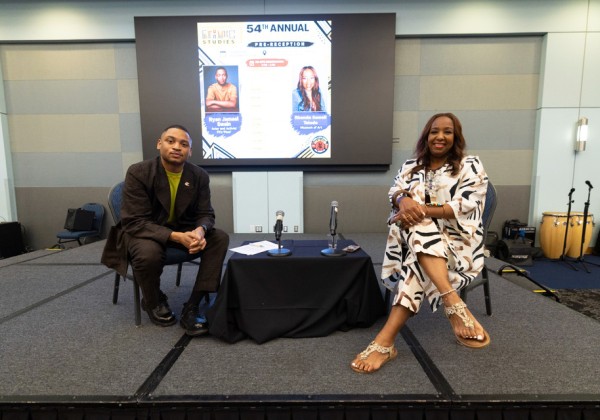 Two guests sit onstage beside a table with microphones during a Cal State LA ethnic studies event.