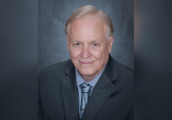 Professional portrait of an individual wearing a gray suit and patterned tie, posed against a dark backdrop. 