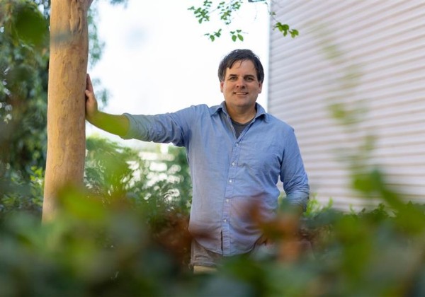 Faculty member stands outdoors on campus, leaning against a tree and smiling, with greenery in the foreground and a building in the background at Cal State LA.