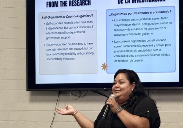 Presenter speaks into a microphone beside a bilingual research presentation during a community feedback meeting.
