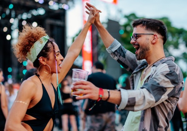 Two people high-five and celebrate while holding drinks in a lively outdoor festival crowd, with a stage and colorful lights visible in the background.