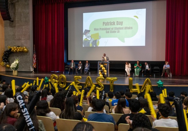 Patrick Day speaks at a podium during the GO East LA celebration as students raise yellow cheering sticks.