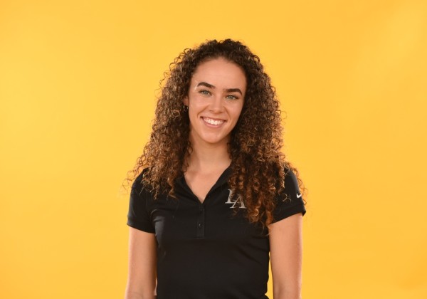Smiling Cal State LA athlete with curly hair in a black polo shirt, standing in front of a bold yellow background.