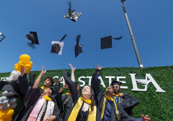 Graduates in black caps and gowns celebrate by tossing their decorated caps into the air, smiling beneath a bright blue sky in front of a Cal State LA backdrop with yellow and gray balloons.