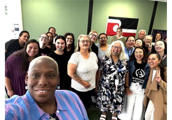A group of smiling people gather for a selfie during a cultural visit, with a red, black, and white flag in the background and a green wall behind them.