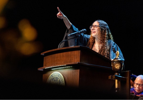 Cal State LA student delivers a commencement speech from a podium, pointing upward with enthusiasm while wearing a graduation gown.