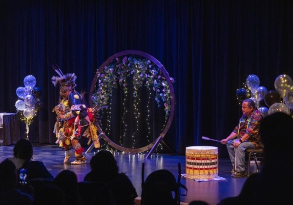 An Indigenous performer dances in traditional regalia on stage during IndigiGrad at Cal State LA, with a drummer, floral ring, and decorative balloons in the background.