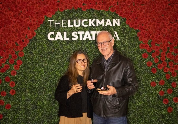 Two guests pose in front of a red rose and greenery photo wall with “The Luckman” and “Cal State LA” signage.