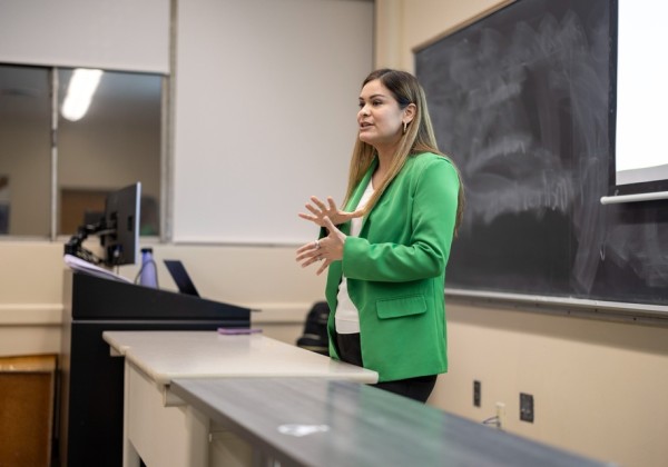 A speaker in a green blazer gestures while presenting at the front of a classroom, standing beside a desk and chalkboard at Cal State LA