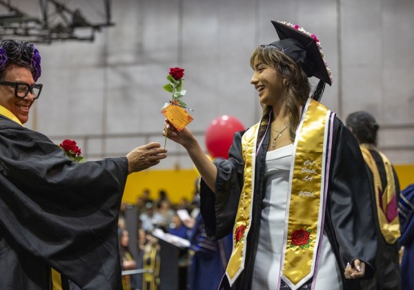 Graduate wearing a decorated cap and gold Cal State LA 2026 stole receives a red rose during a Nuestra Graduación celebration.
