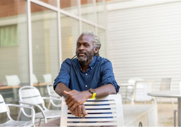 Portrait of Olaseni Sode seated in an outdoor campus courtyard at Cal State LA, resting arms on the back of a white metal chair, with tables and glass walls in the background.