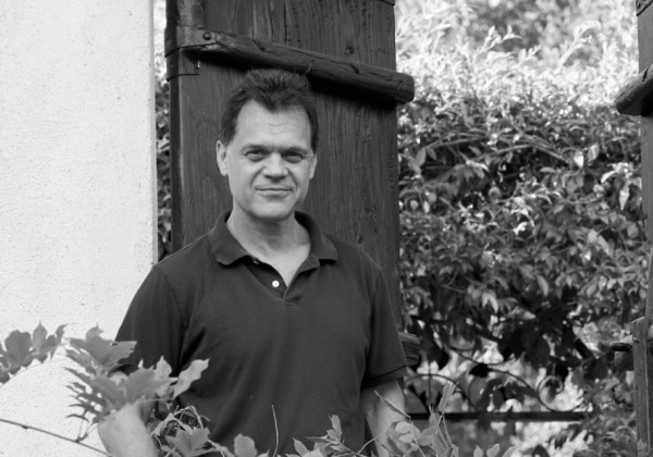 Black-and-white portrait of a man standing beside a wooden gate and stucco wall, surrounded by foliage.