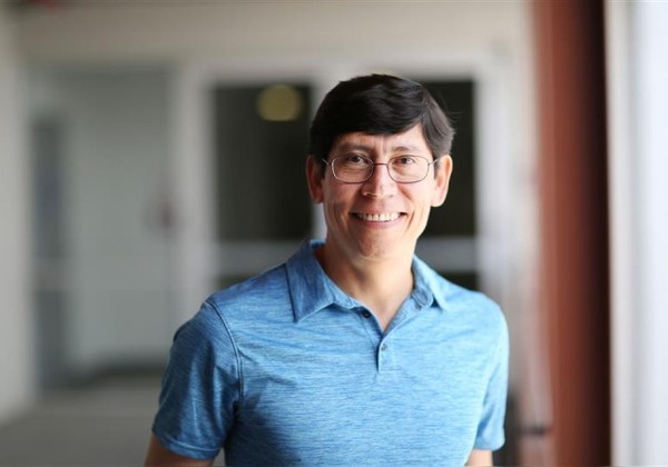 Portrait of a smiling man in a blue polo shirt and glasses standing in a softly lit indoor corridor with a blurred background.