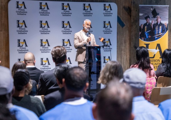 A speaker addresses attendees from a podium in front of a Cal State LA Prison Graduation Initiative backdrop.