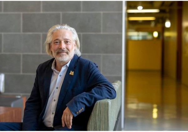 Portrait of Stephen Trzaskoma seated on a bench inside a modern campus building at Cal State LA, with tiled walls and a hallway in the background.