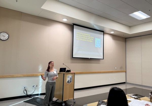 A Cal State LA student presents research findings at a symposium, standing beside a screen displaying their project title.