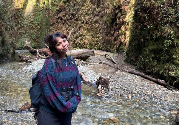 Student wearing a backpack and binoculars stands in a rocky stream during outdoor fieldwork in a forested canyon.