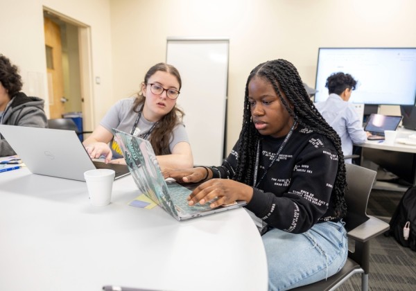 Students collaborate on laptops during a hands-on coding workshop at Cal State LA, seated around a table in a modern classroom with whiteboards and screens.