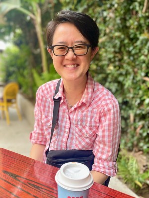 Cynthia Wang smiles while seated outdoors at a café, wearing black glasses and a pink-and-white plaid shirt. A takeaway coffee cup sits on the red wooden table in front of her, with lush greenery in the background.
