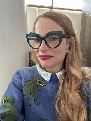 Woman with long hair, green glasses, big bird earrings and a dinosaur sweater.