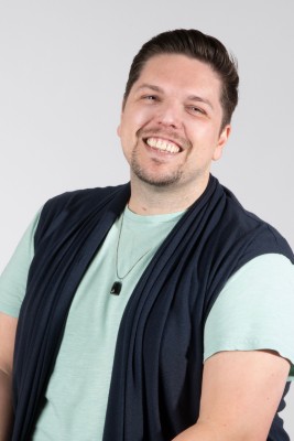 Photo of a smiling white man with a goatee wearing a light teal shirt, dark blue vest, and black necklace