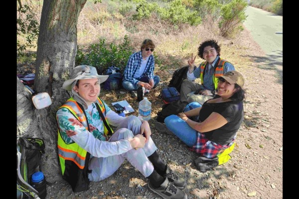 Three students sitting on the ground next to the trunk of the tree smiling.