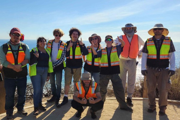 Group of students wearing safety vests and hats in front of the coast on a sunny day.