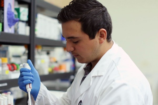 Image of the side profile of a student in a while lab coat and blue gloves using a lab instrument. 