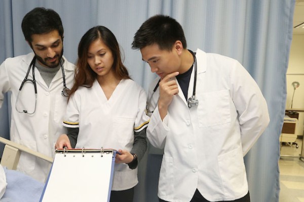 Image of three students in lab coats standing over an educational dummy. 