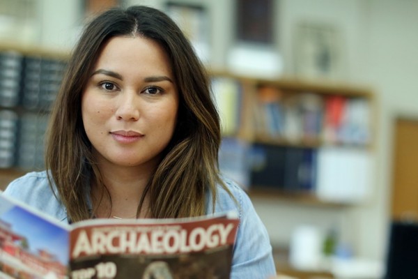 Image of a female student looking directly into the camera as she holds a copy of Archaeology Magazine in a classroom. 