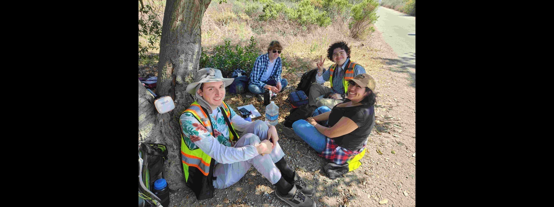 Three students sitting on the ground next to the trunk of the tree smiling.