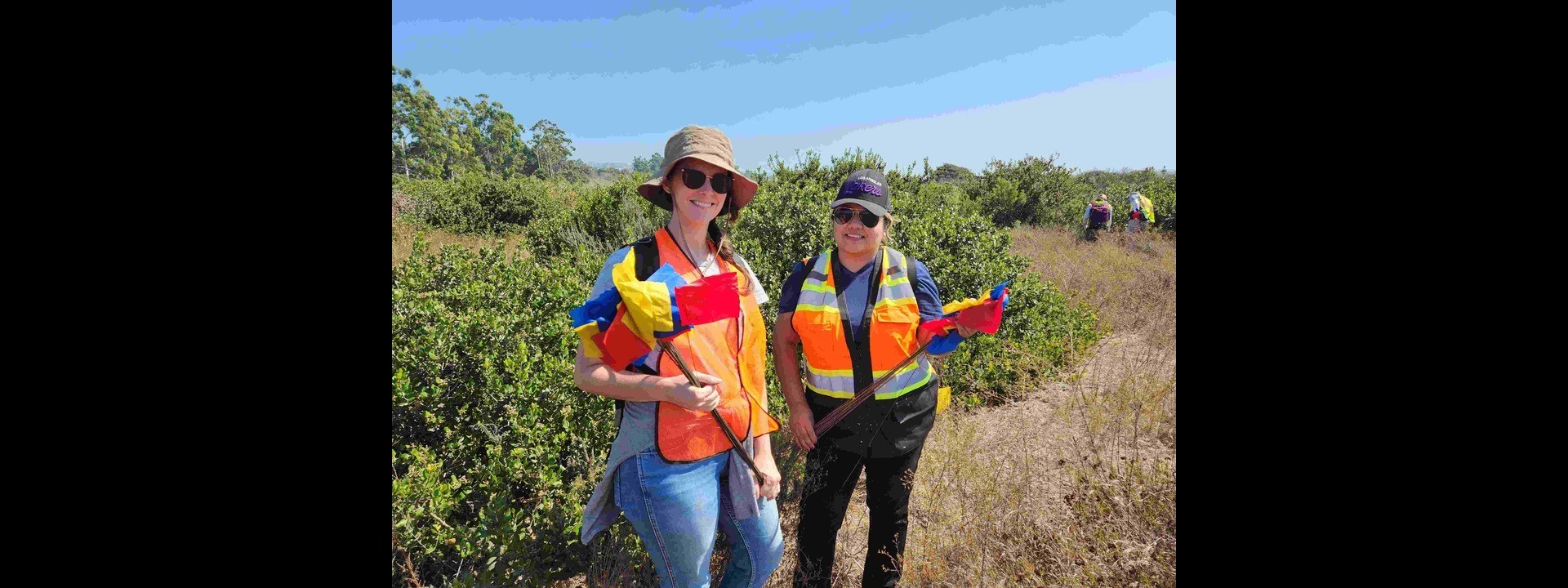 Two students wearing hats holding red and yellow flags smiling for the camera in the field.