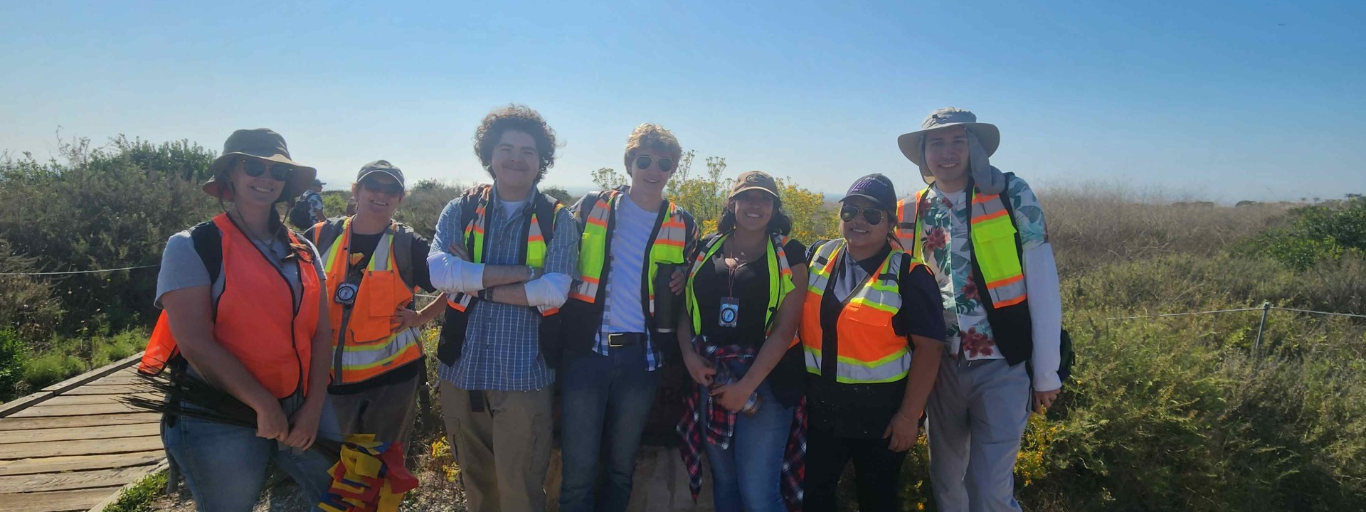 Group of students wearing safety vests and hats in front of the coast on a sunny day.