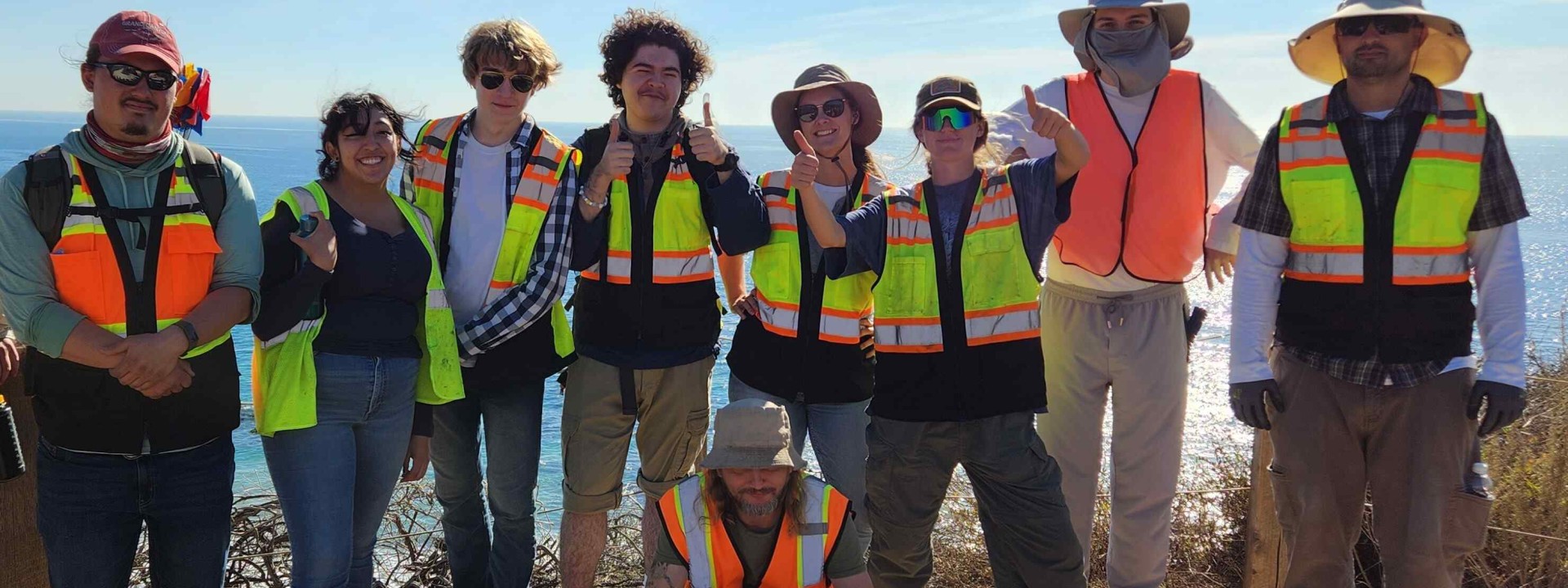Group of students wearing safety vests and hats in front of the coast on a sunny day.