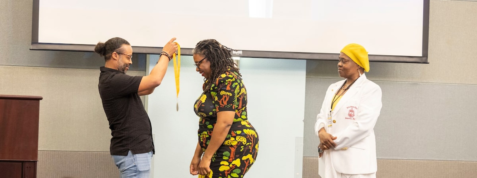 Male thesis mentor donning medal onto female student in a black dress with sunflowers