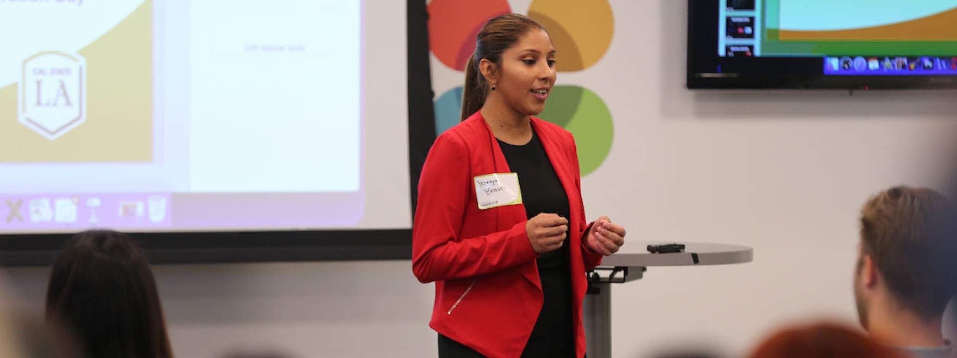 Image of a female student in a red blazer giving a presentation to her classmates. 