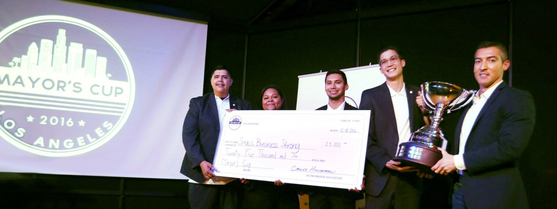 Group of Cal State LA students in business attire on a stage holding a large check near a screen that says "2016 Mayor's Cup"