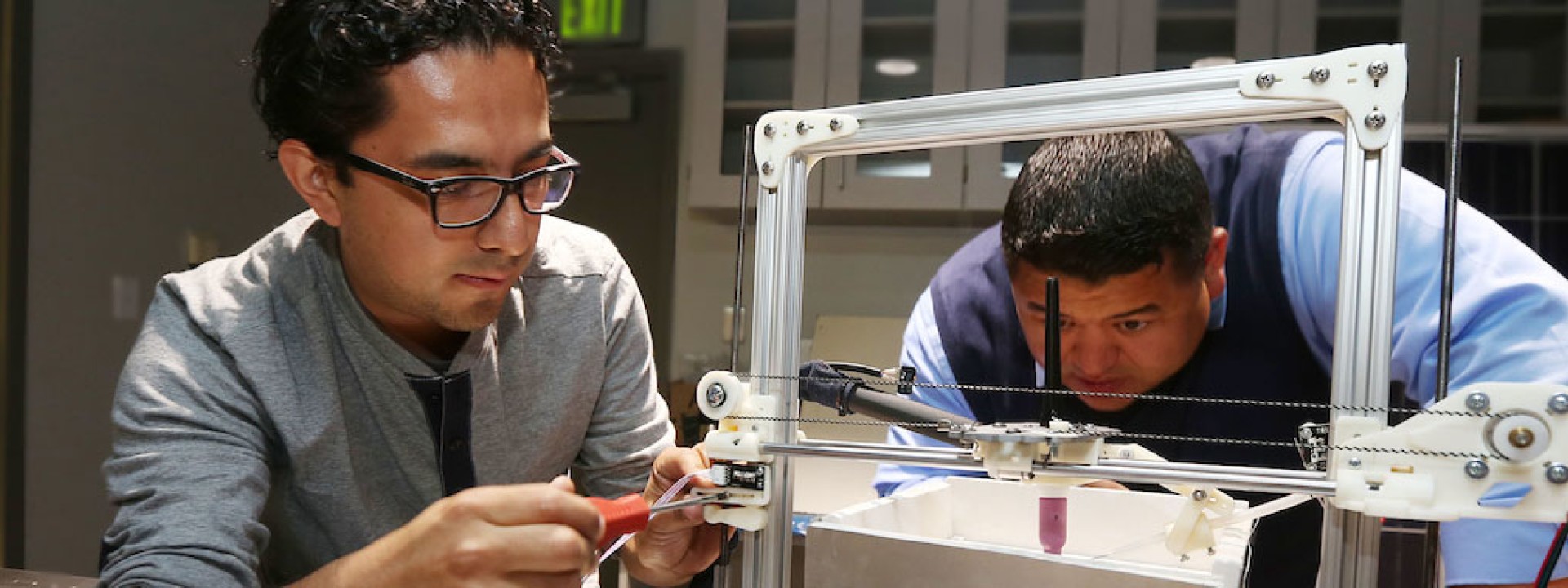 Image of two engineering students looking at a metal frame device intently. 