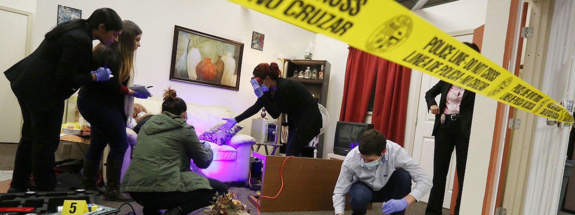 Image of a mock crime scene with caution tape and black lights, with forensic students investigating. 